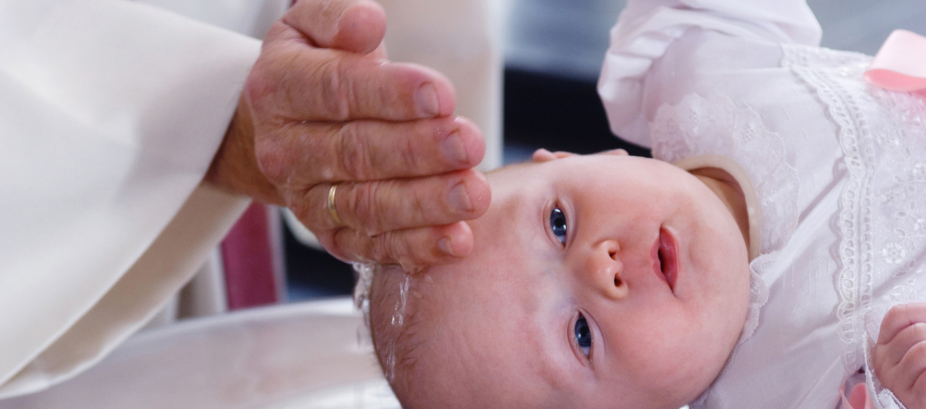 Nærbilde av en baby iført hvit dåpskjole som får vann helt over hodet under en dåpsseremoni.